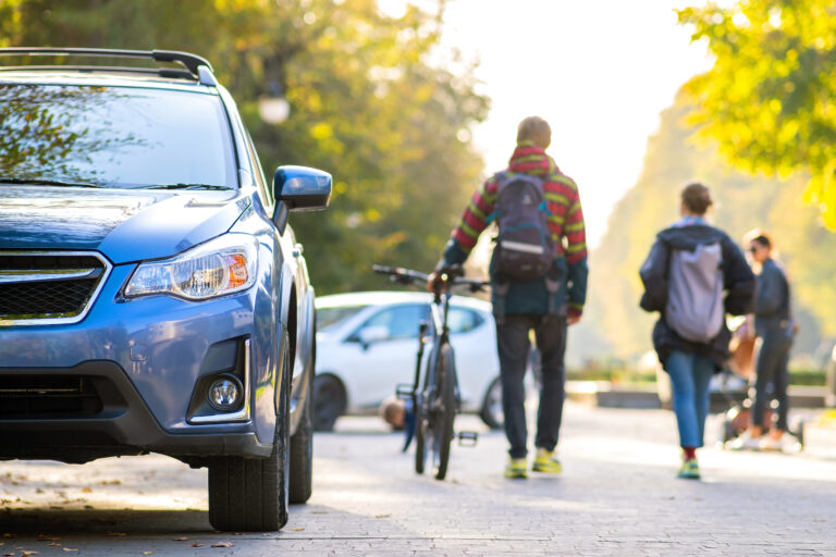 Blue car in the forefront, peoples walking in the background on the street.